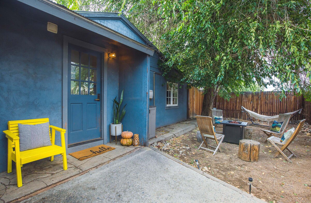 A private outdoor space features a blue exterior with a welcoming entrance. A bright yellow bench sits next to the doorway. Seating is arranged around a fire pit, complemented by a hammock hanging between trees, creating a cozy atmosphere for relaxation.