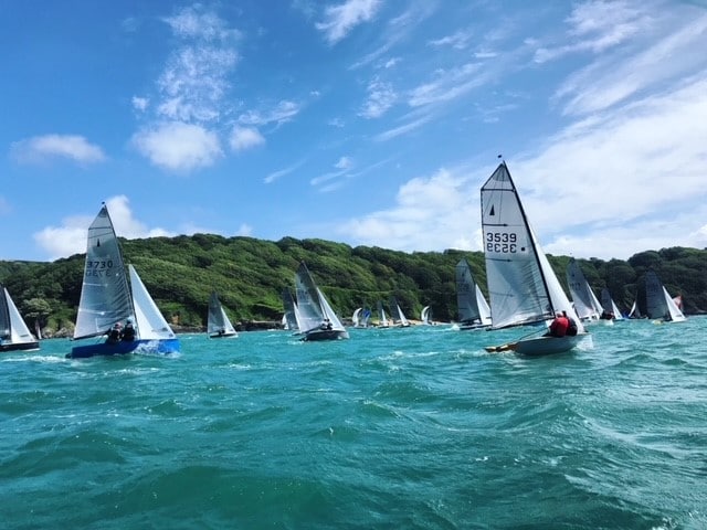 A lively scene on the water showcases numerous sailboats gliding across turquoise waves under a clear blue sky. Lush greenery dots the background, creating a vibrant contrast against the sea and enhancing the sense of movement and activity.
