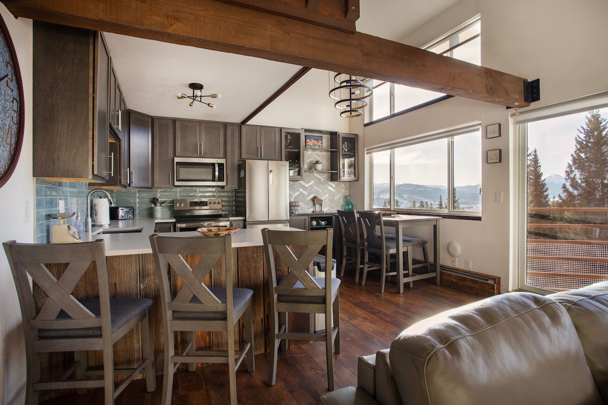 A modern kitchen area is highlighted, featuring dark cabinetry and stainless steel appliances. A central island offers seating for six, complemented by a dining area with mountain views through large windows. Wooden beams accentuate the space, creating an open and inviting atmosphere.