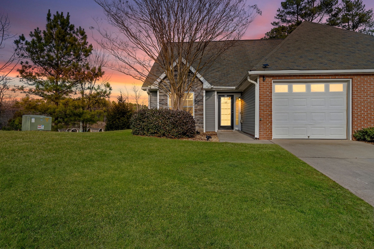 The exterior of the single-level ranch home is shown at dusk, with soft lighting illuminating the front entrance. The manicured lawn surrounds the house, while trees and a colorful sunset can be seen in the background.
