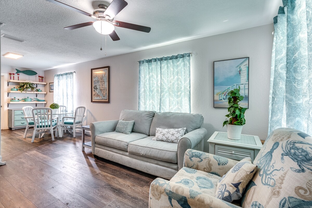 A living area features a light blue sofa and an armchair with a nautical design. Natural light enters through two windows adorned with sheer curtains. A dining table with white chairs is visible in the background, with decorative shelving displaying plants and artwork.