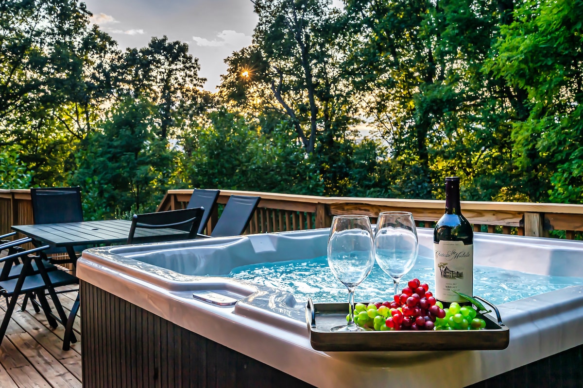 A spacious hot tub is situated on a wooden deck, surrounded by lush trees. A tray holds two glasses and a bottle of wine alongside fresh grapes, inviting relaxation. The evening sky is visible in the background, showcasing a blend of colors as the sun sets.