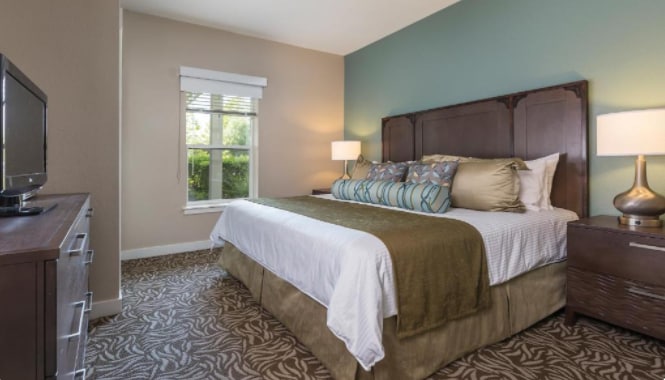 A spacious bedroom features a king-sized bed adorned with decorative pillows and a soft throw blanket. Natural light filters through a window with blinds, illuminating the room's soothing green wall and stylish furnishings, including a dresser and bedside lamps.