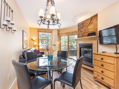 A dining area features a round glass table surrounded by four black chairs. A wooden dresser sits nearby, and a wall-mounted TV is above a gas fireplace set within a stone surround. Large windows allow natural light and views of the surrounding trees.