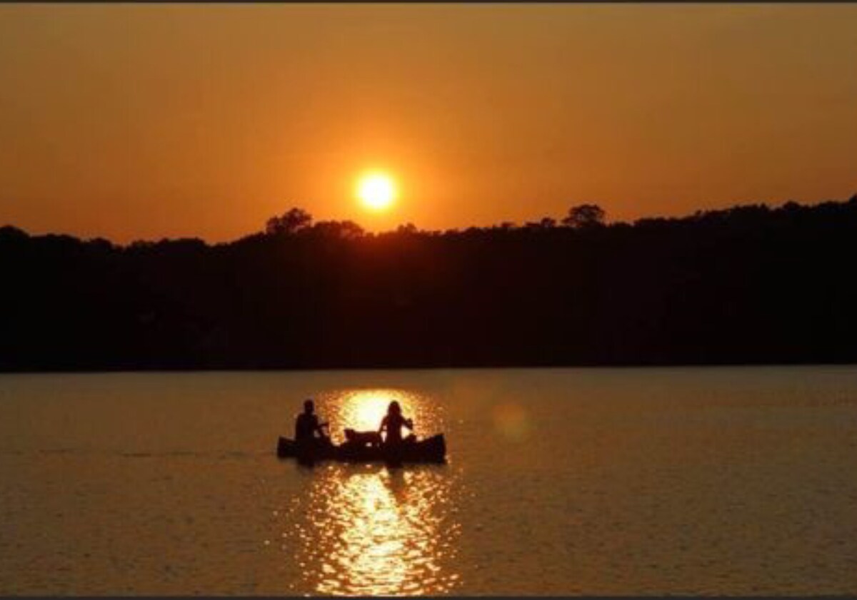 A serene sunset reflects on the calm waters of the lake, where two individuals are seen paddling in a canoe. The golden hues of the sun create a warm glow, while trees outline the horizon, enhancing the tranquil atmosphere.