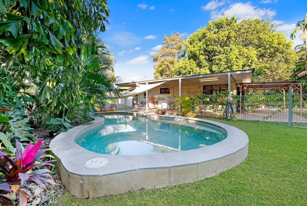 A lagoon-style swimming pool is set within a manicured garden, surrounded by tropical plants and greenery. The pool's smooth surface reflects the blue sky and is bordered by a spacious grassy area, providing an inviting outdoor space for relaxation and leisure.