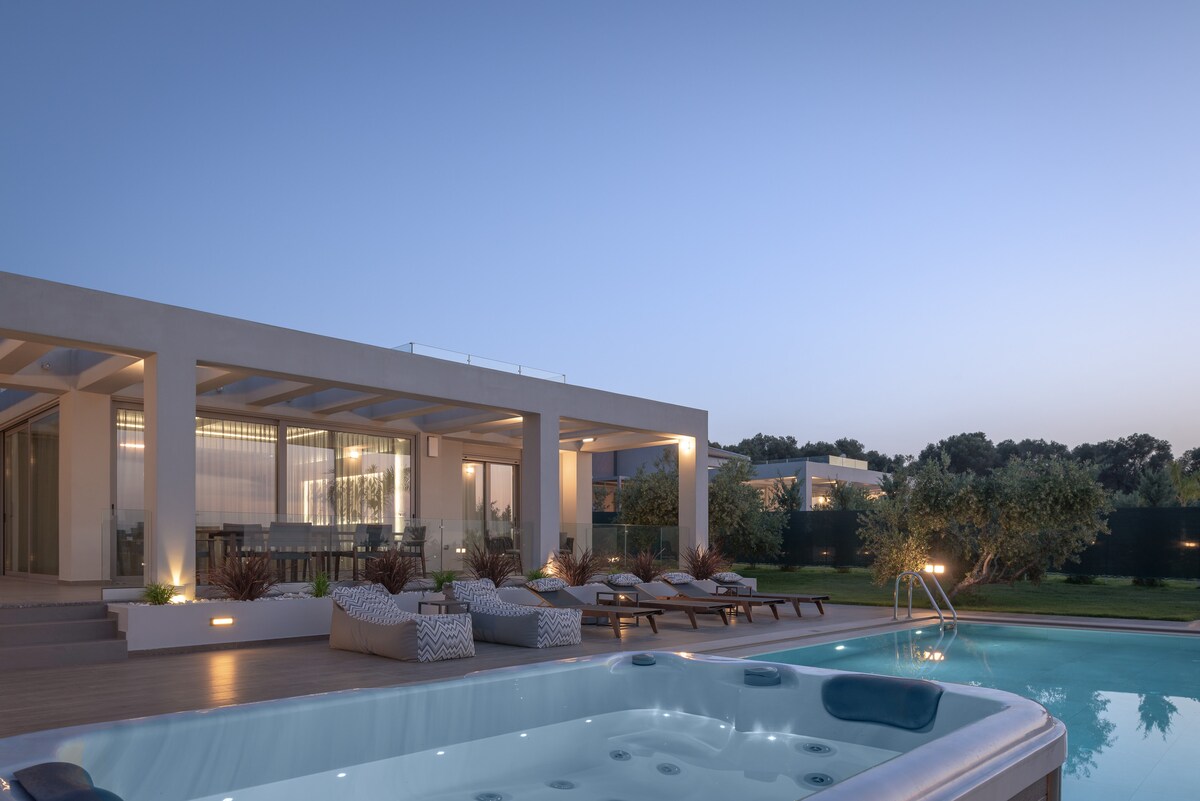 An outdoor space features a private pool and heated spa whirlpool, surrounded by loungers and shaded seating. The villa's sleek design is accentuated by warm outdoor lighting as the sky transitions to dusk, creating a serene environment for relaxation.