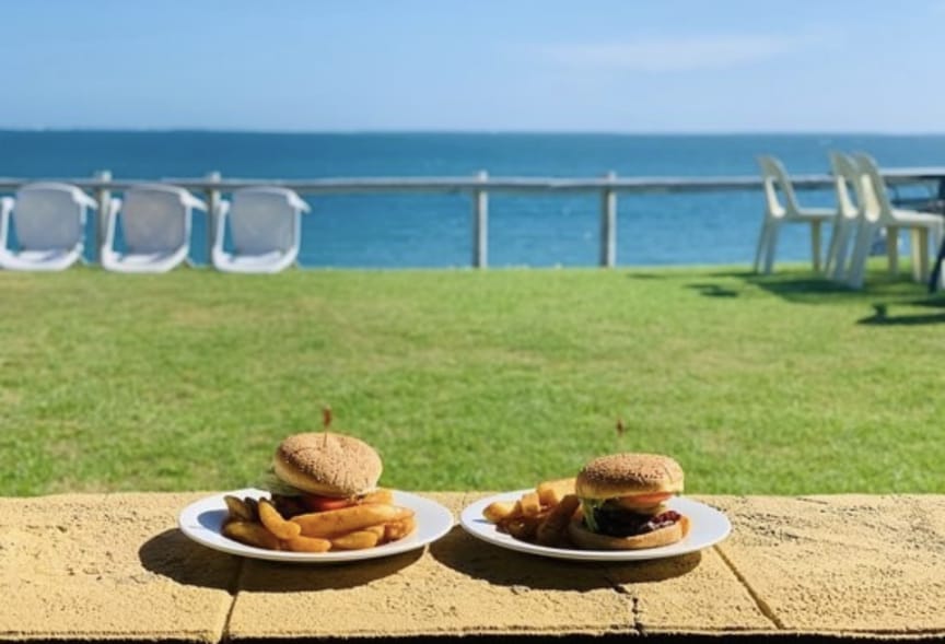 Two plates of freshly prepared burgers and fries are displayed on a stone ledge, with a panoramic ocean view in the background. A grassy area is present in the foreground, complemented by lounge chairs along the water's edge.