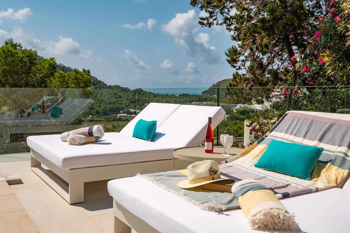Two spacious sun loungers are positioned on a terrace, offering a clear view of the landscape and distant sea. Bright towels and a hat rest on the loungers. A small table holds a bottle of wine and glasses, inviting relaxation in a serene outdoor setting.