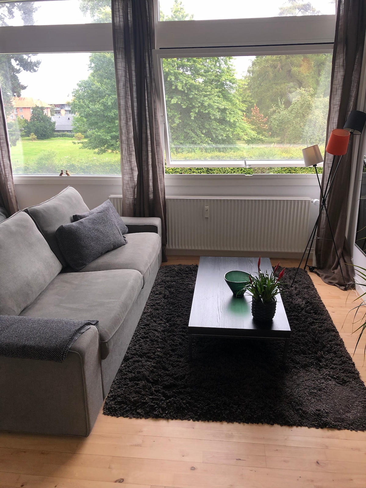 A comfortable living area features a soft gray sofa adorned with a textured cushion. A small black table sits in front, accompanied by a potted plant. Large windows allow natural light to illuminate the space, providing a view of greenery outside.