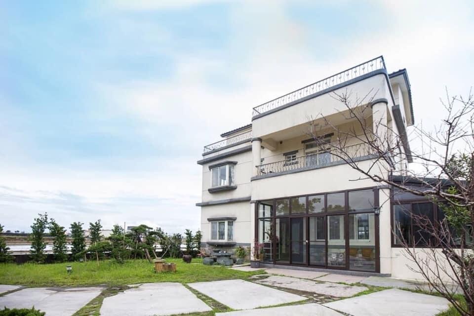 A two-story building showcases a modern design with large glass windows and a spacious outdoor area. Lush greenery surrounds the property, creating a serene atmosphere. A paved pathway leads to the entrance, enhancing the welcoming feel of the residence.