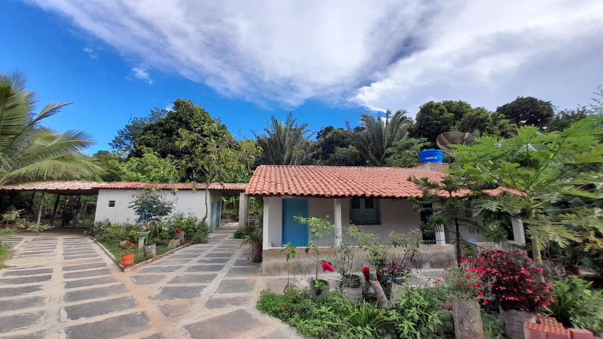 A charming single-story structure features a rustic clay tile roof and light-colored walls. Lush greenery and flowering plants surround the home, while a wide stone pathway guides visitors through the garden area, leading to the entrance.