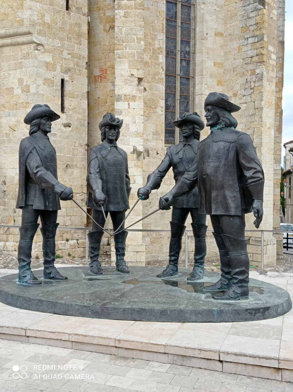 A bronze statue depicts four historical figures engaged in a duel, standing on a circular stone platform. The figures are captured mid-action, with swords drawn, displaying intricate details in their clothing and expressions. A stone wall is visible in the background.