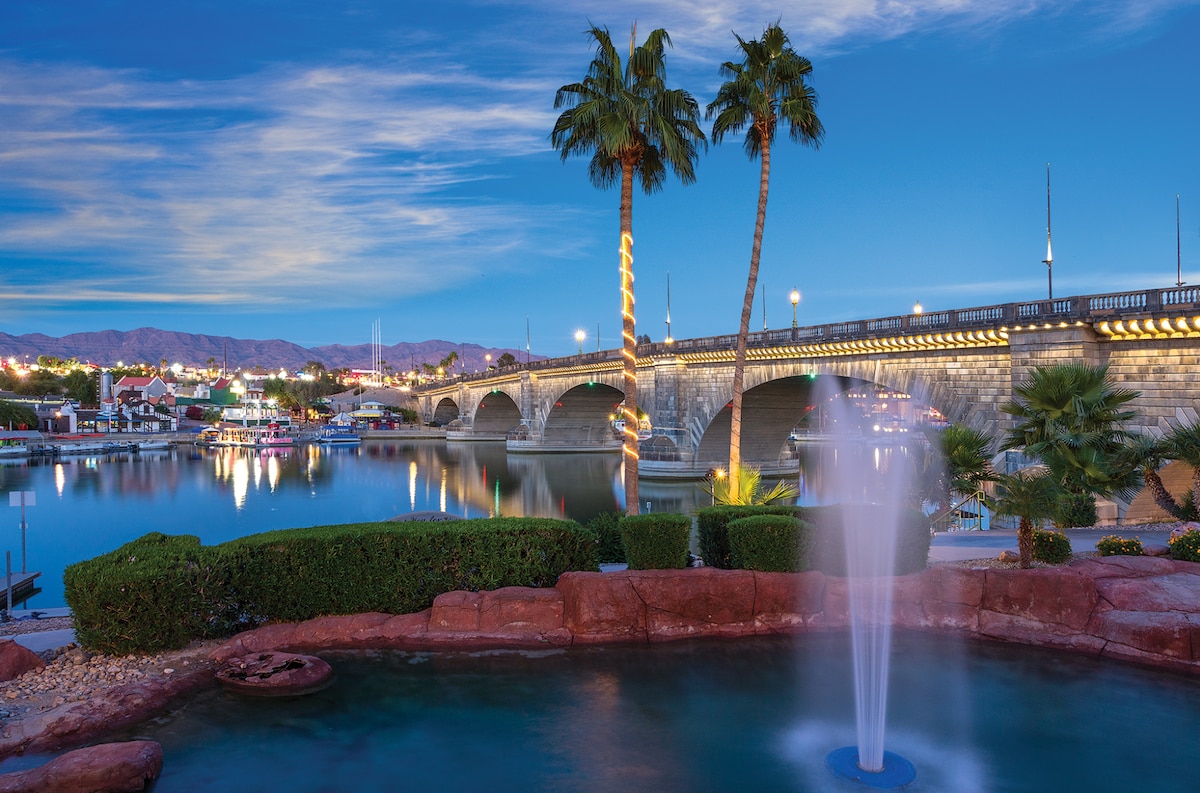 A serene waterfront scene showcases a fountain surrounded by lush greenery, with palm trees lining the shore. In the background, a stone bridge spans the river, illuminated by soft lights, while distant mountains are bathed in a gentle twilight glow.