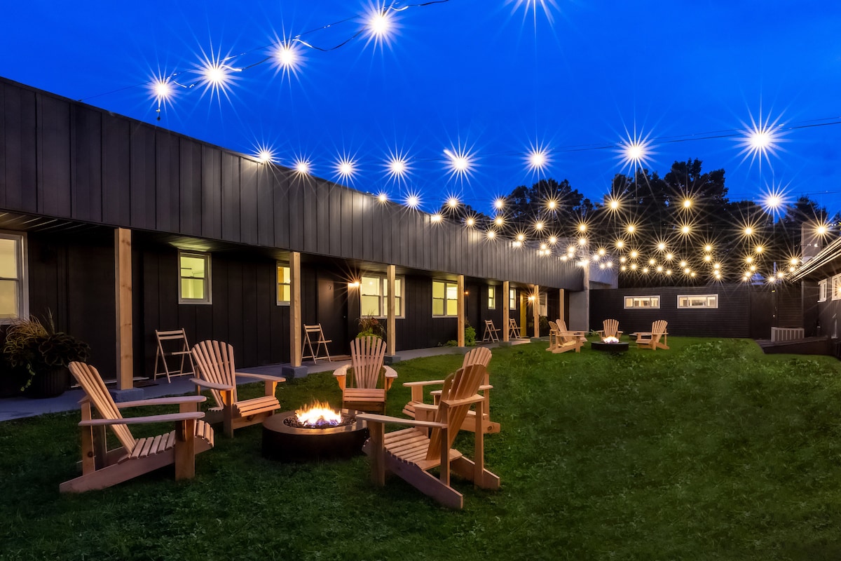 An outdoor area is illuminated by string lights, creating a warm ambiance. Several wooden Adirondack chairs are arranged around a central fire pit. The grassy space is bordered by a low building, featuring large windows and a welcoming exterior.