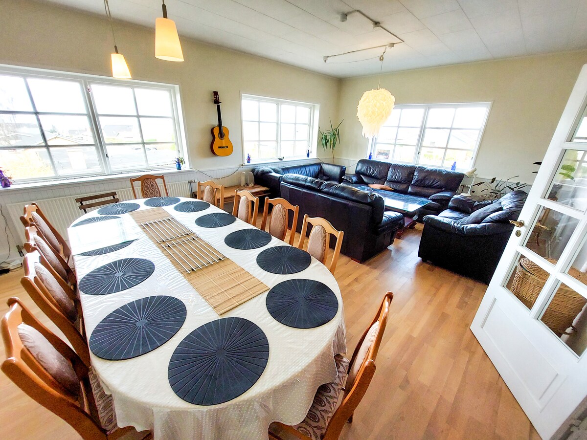 A spacious dining area is featured, with a large table set for twelve, adorned with dark circular placemats. Comfortable seating is arranged around the room, including a sectional sofa. Large windows allow natural light to fill the space, creating an open and inviting atmosphere.