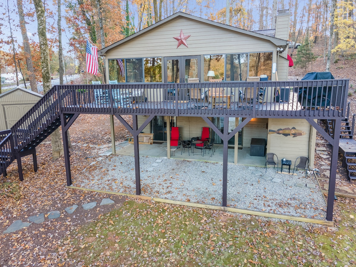 A wooden deck extends from the cozy cabin, bordered by trees displaying autumn leaves. Below, a gravel area is furnished with a seating arrangement, including red chairs. An American flag and a star accent the exterior, contributing to a welcoming outdoor space.