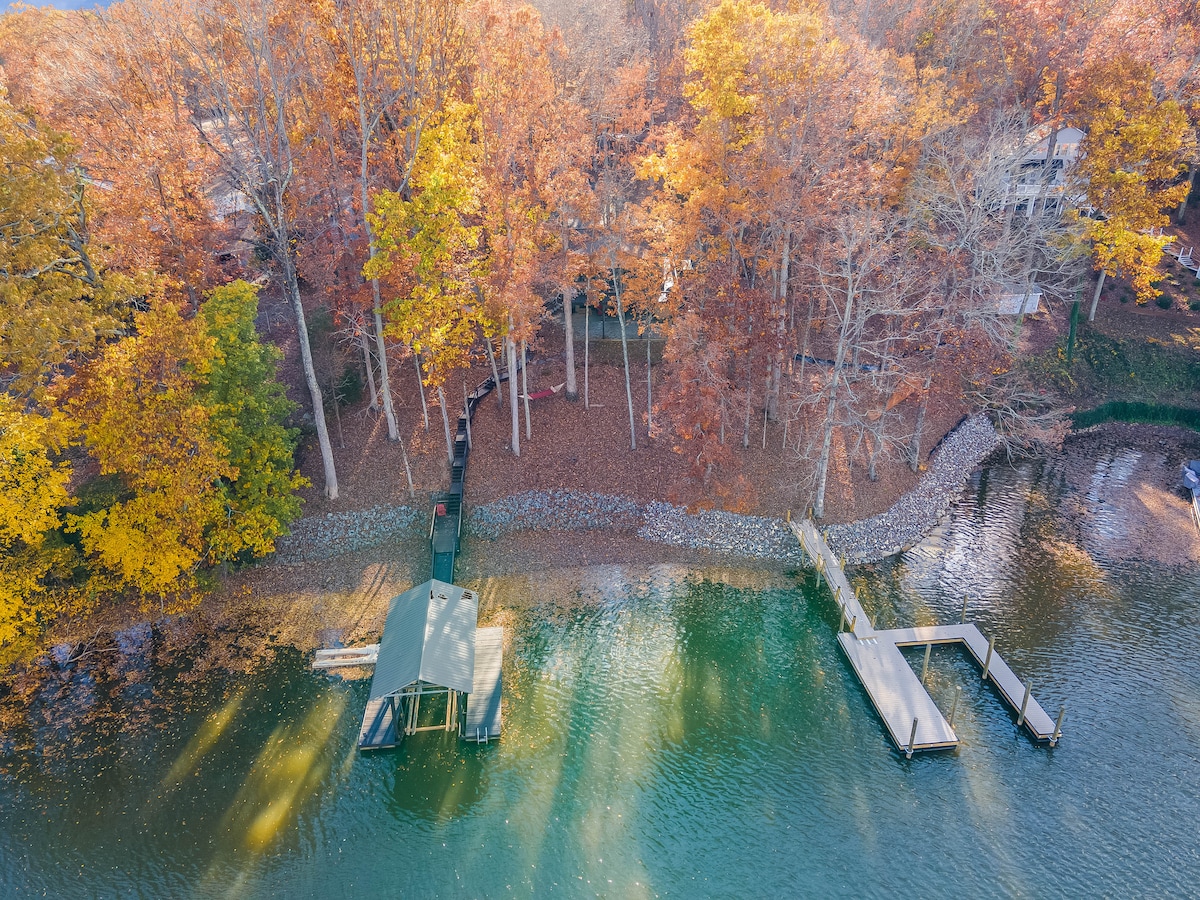 Vibrant autumn foliage surrounds a tranquil waterway, with a private dock accessible for boat mooring. The brown and orange leaves contrast beautifully against the clear blue water. A serene, wooded area offers a peaceful setting, reflecting the calm atmosphere of the cove.