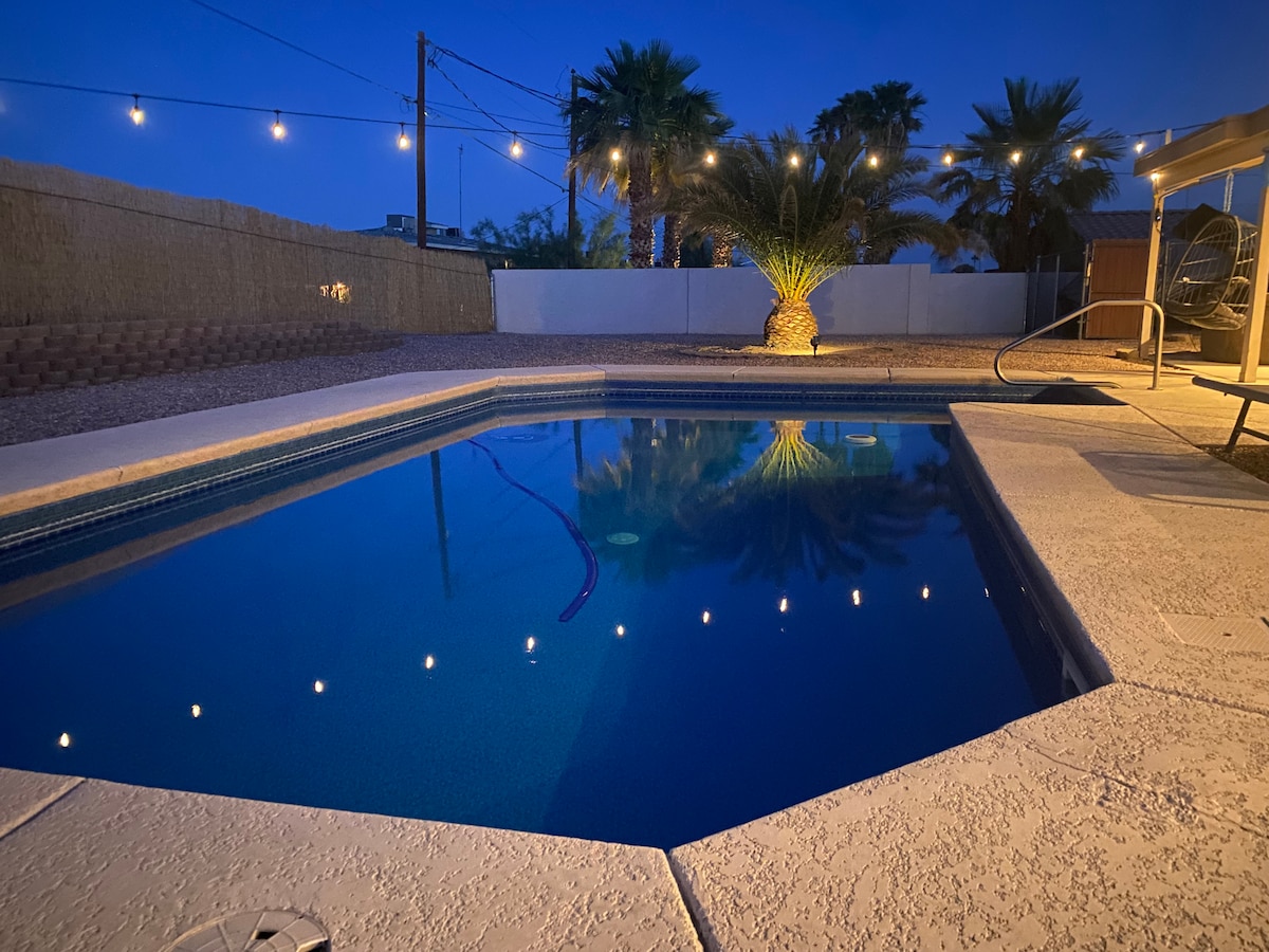 A tranquil pool area is depicted at night, with string lights illuminating the space. The deep blue water reflects the surrounding palm trees and sky, while a nearby lounging chair adds to the ambiance. The outdoor setting offers a peaceful retreat.