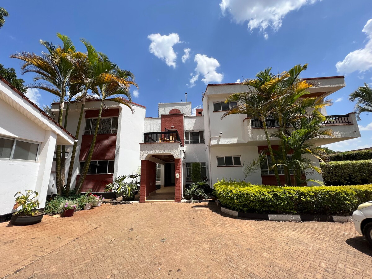The exterior of the building is showcased, featuring a two-story design with a central entrance framed by lush greenery. Palm trees add to the serene ambiance, while a paved driveway leads to the front door. The blue sky and scattered clouds enhance the inviting setting.
