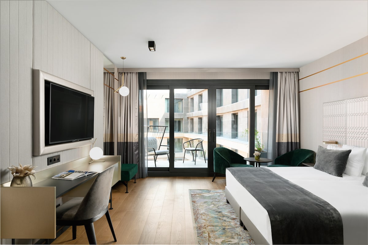A modern guest room features a spacious bed with grey bedding and a striped area rug. Large glass doors open to a balcony with outdoor seating, allowing natural light to fill the room. A TV and desk are positioned against the wall, enhancing functionality.