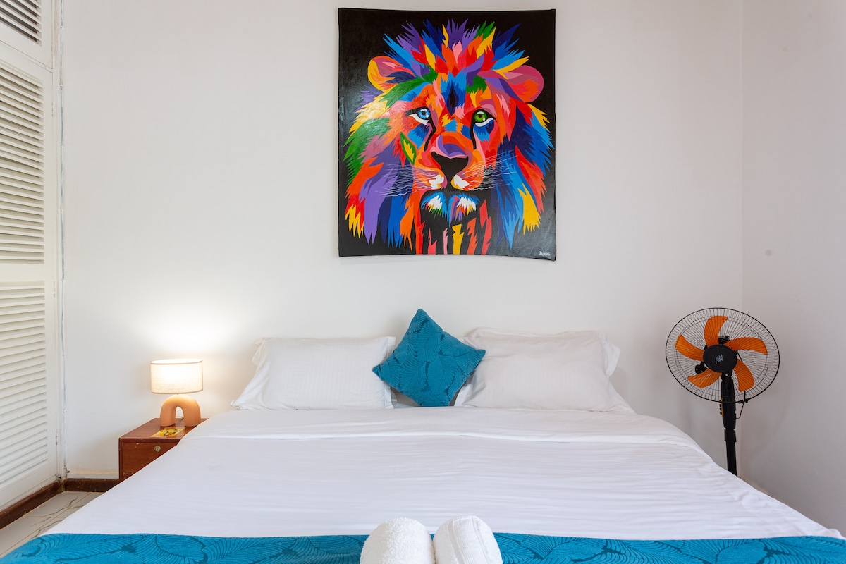 A bright bedroom features a spacious bed with crisp white linens and a colorful lion painting above the headboard. A blue decorative pillow adds a pop of color, while a bedside lamp and a standing fan are positioned nearby, contributing to a pleasant and functional space.
