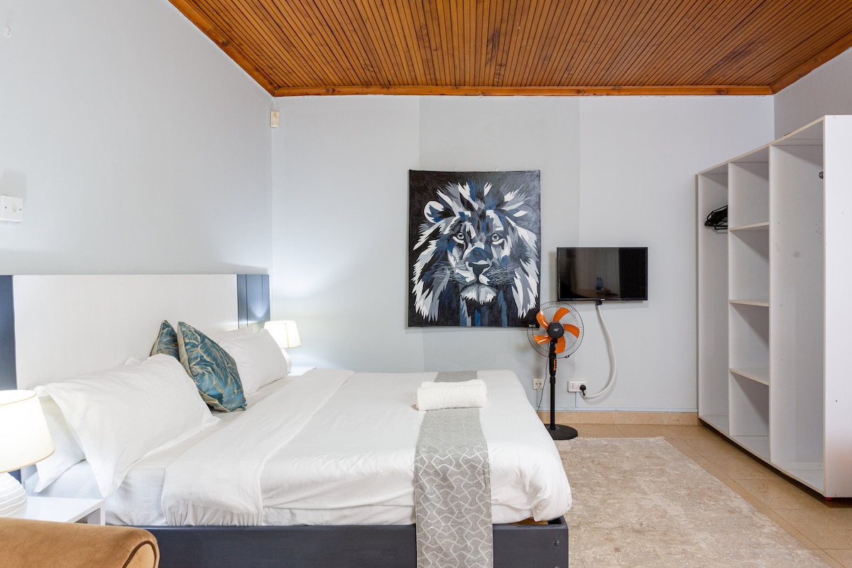 A spacious sleeping area is featured with a large bed adorned with crisp linens and decorative pillows. The wall displays a striking lion artwork, while a flat-screen TV is mounted nearby. Illuminated bedside tables enhance the room's functionality.