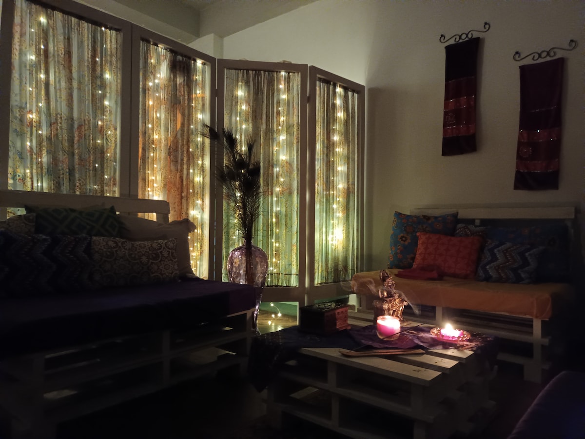 A cozy sitting area is surrounded by screens draped in shimmering lights, enhancing the serene atmosphere. Colorful cushions are arranged on wooden pallet seating, and candles cast a gentle glow on the space, inviting relaxation and contemplation.