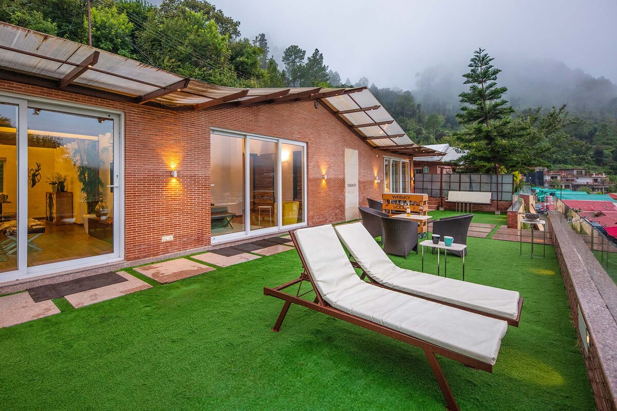 The outdoor deck showcases sun loungers arranged on a lush green lawn, with a terrace equipped for entertaining. The backdrop features mist-covered mountains, with modern brick walls of the chalet illuminated by soft lighting, creating a serene environment.