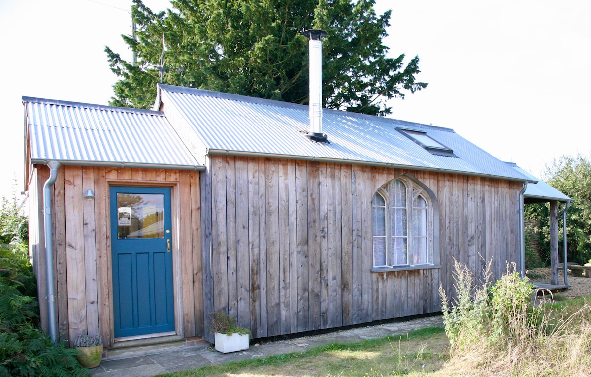 A timber-framed building features a sloped metal roof and a welcoming blue door. An arched window adds character to the exterior, while a stone pathway leads to the entrance. Natural greenery surrounds the structure, contributing to a peaceful setting.