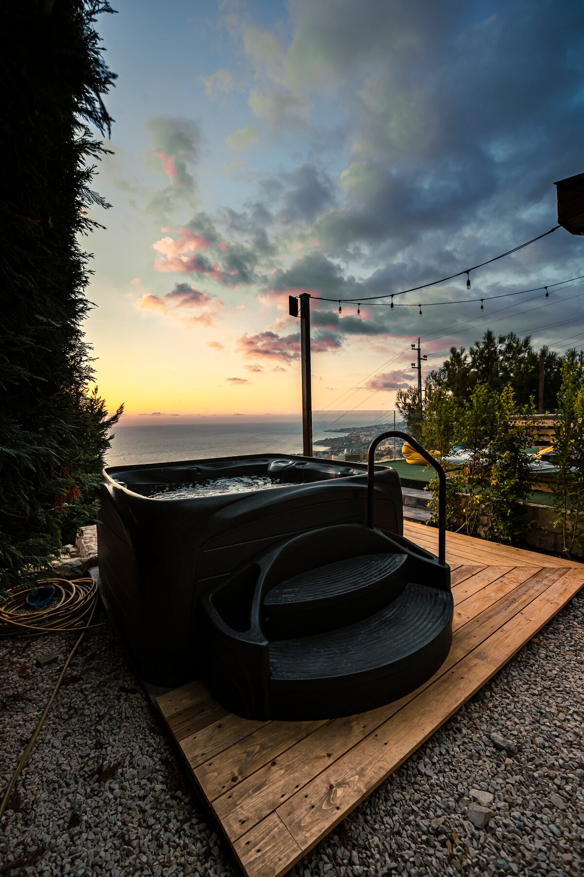 A private hot tub is situated on a wooden deck, surrounded by natural greenery. The sunset casts a warm glow over the scene, with clouds painted in soft hues. String lights are visible overhead, enhancing the tranquil outdoor atmosphere.