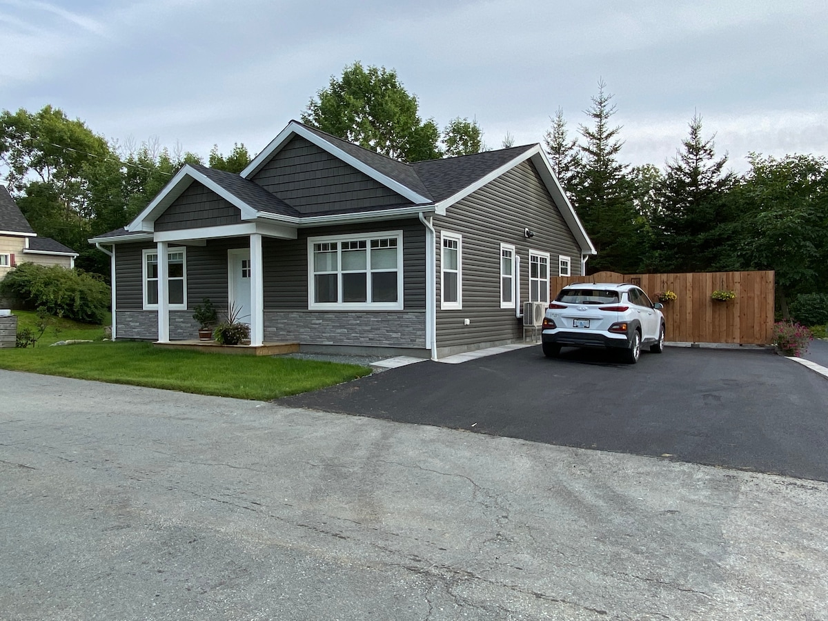 A newly constructed bungalow features a modern exterior with gray siding and stone accents. A paved driveway provides parking for vehicles, and a well-maintained lawn adds to the welcoming appearance. Large windows allow natural light into the home, enhancing the inviting facade.