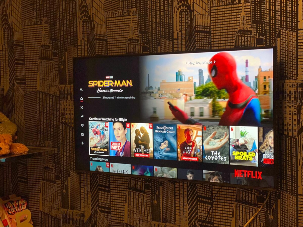 A smart TV is mounted on a decorative patterned wall, displaying a popular streaming service interface. A colorful assortment of movie titles is visible, including 'Spider-Man: Homecoming,' along with various recommended titles arranged below. The overall ambiance reflects a modern entertainment setup.