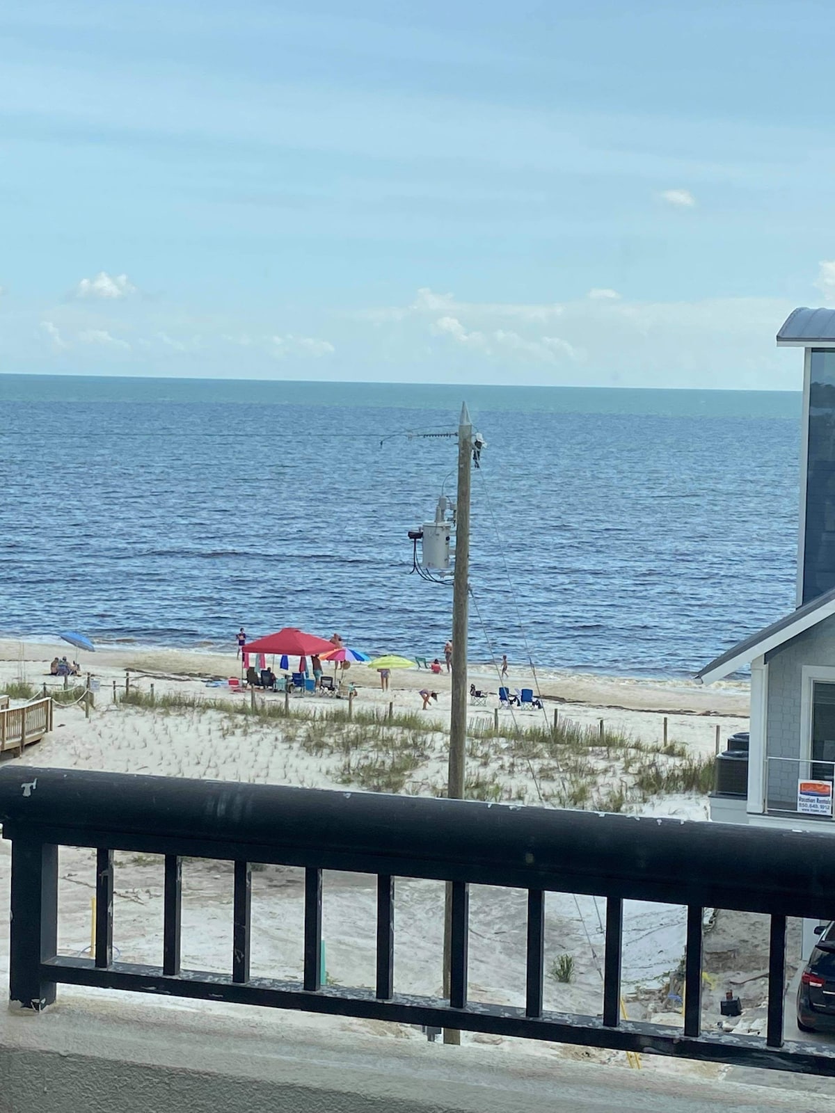 A view overlooking the beach reveals gentle waves and a clear sky. A red and white canopy is set up on the sand, with people enjoying the shoreline. The scene is framed by a black balcony railing and nearby beach grass.