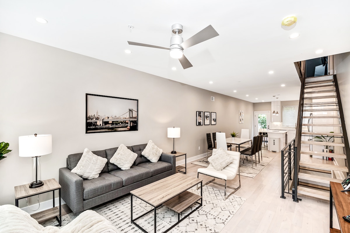 An open concept living area is showcased, featuring a comfortable grey sofa with accent pillows and a matching armchair. A wooden coffee table sits in front, accompanied by two modern lamps. Natural light floods the space, which includes stairs leading to the upper floor in the background.