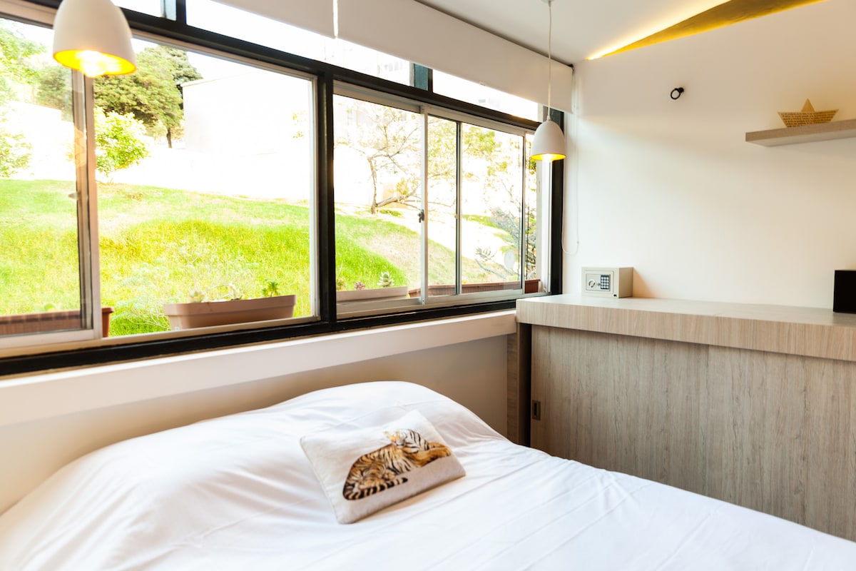 A bright sleeping area is featured, with large windows allowing natural light to illuminate the space. A spacious bed is present, topped with a simple decorative pillow. A wooden shelf is positioned against the wall, and greenery can be seen outside through the windows.