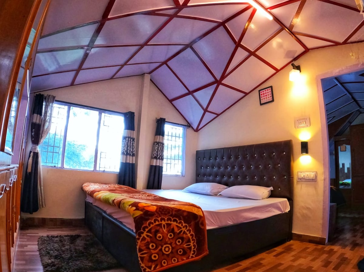 A family suite features a spacious bed adorned with a patterned blanket. Large windows allow natural light to fill the room, showcasing a cozy atmosphere. The ceiling is designed with a unique geometric pattern, and the wooden flooring adds warmth to the space.