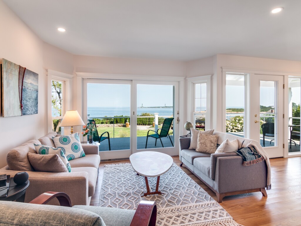A comfortable living area is designed with large windows offering views of the ocean and distant lighthouses. Two sofas are positioned around a coffee table, with a patterned area rug beneath. Soft lighting and coastal artwork enhance the serene environment.