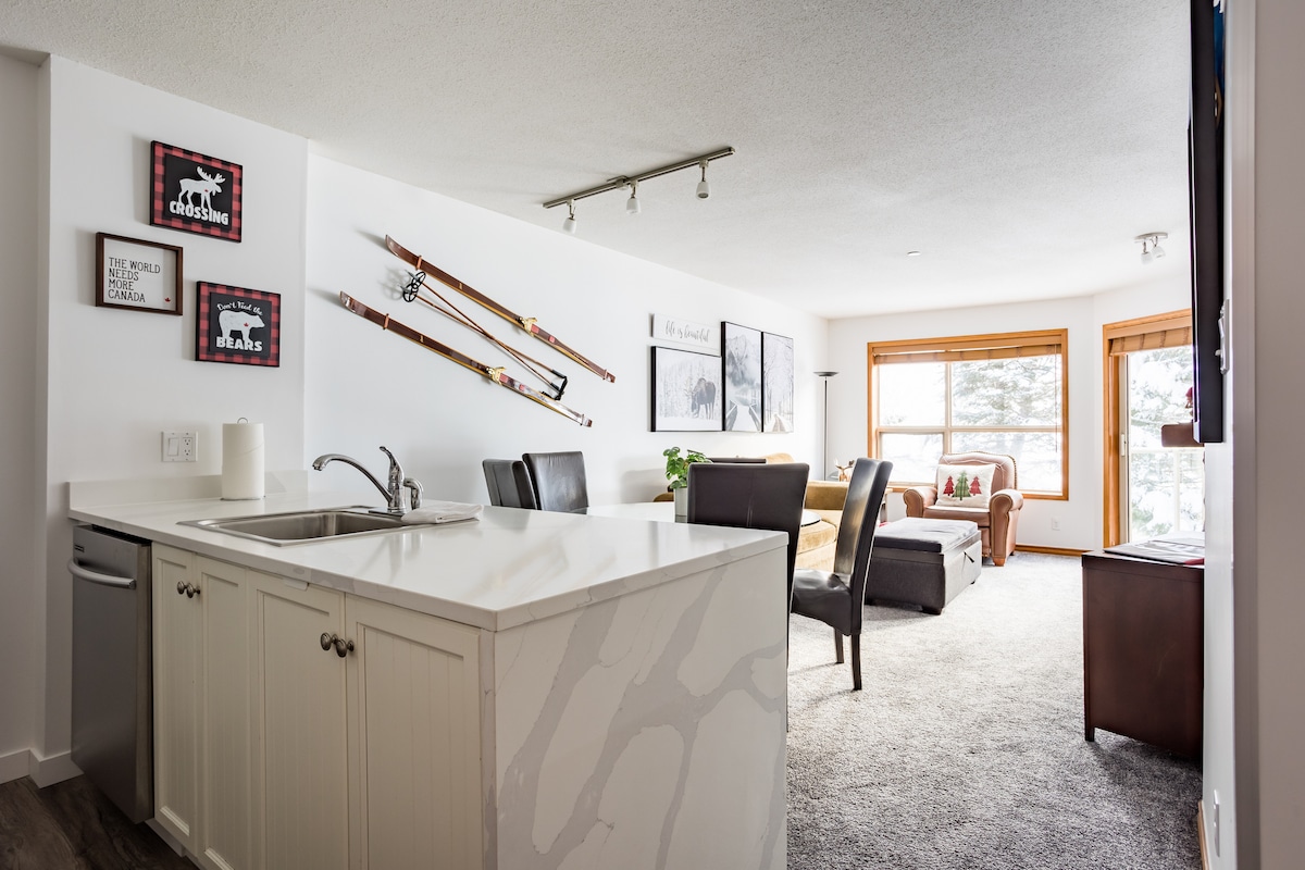 An open-plan living space is visible, featuring a modern kitchen with a sleek countertop and stainless-steel appliances. Decorated with framed artwork and vintage skis on the wall, the cozy seating area is illuminated by natural light from large windows, highlighting the inviting carpeted floor.