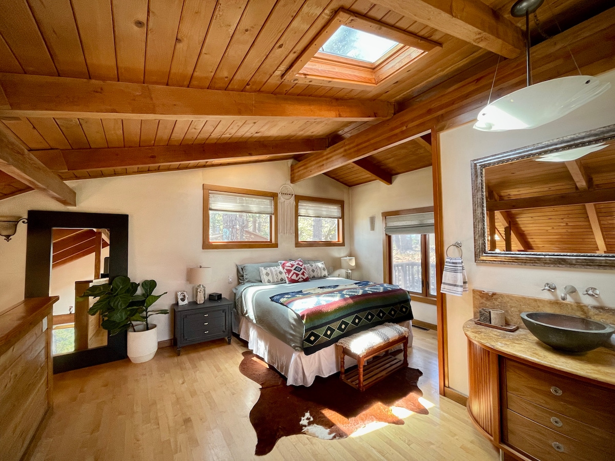 A warmly lit bedroom features wooden beams and a skylight above a spacious bed. A cozy patterned blanket is draped over the bed, complemented by decorative pillows. A stylish mirror and indoor plant enhance the natural aesthetic, while wooden flooring adds warmth to the space.