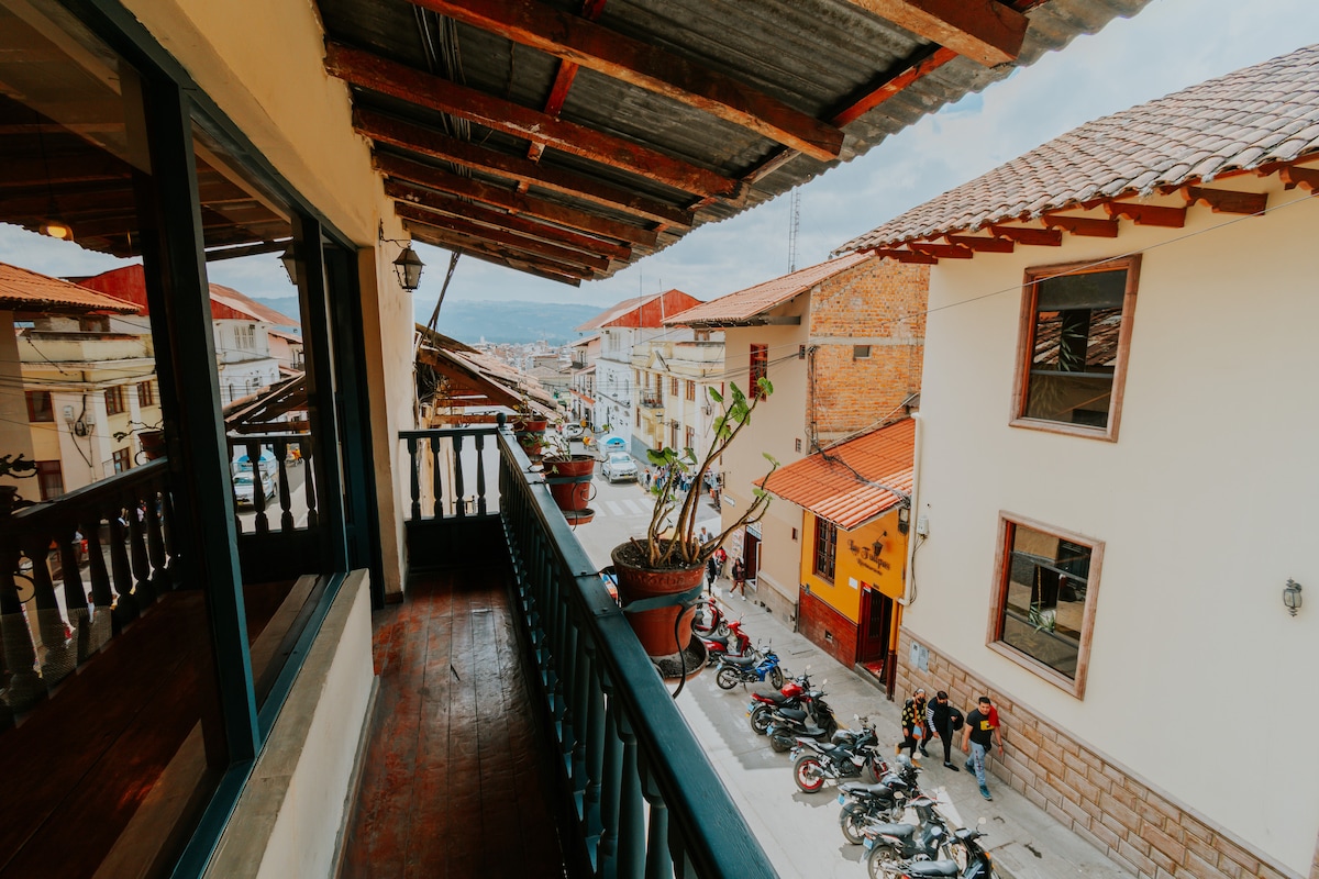 A spacious balcony offers a view of the bustling street below, lined with colorful buildings and parked motorcycles. Potted plants provide greenery, and the wooden railing frames the scene, enhancing the inviting atmosphere of this central location.