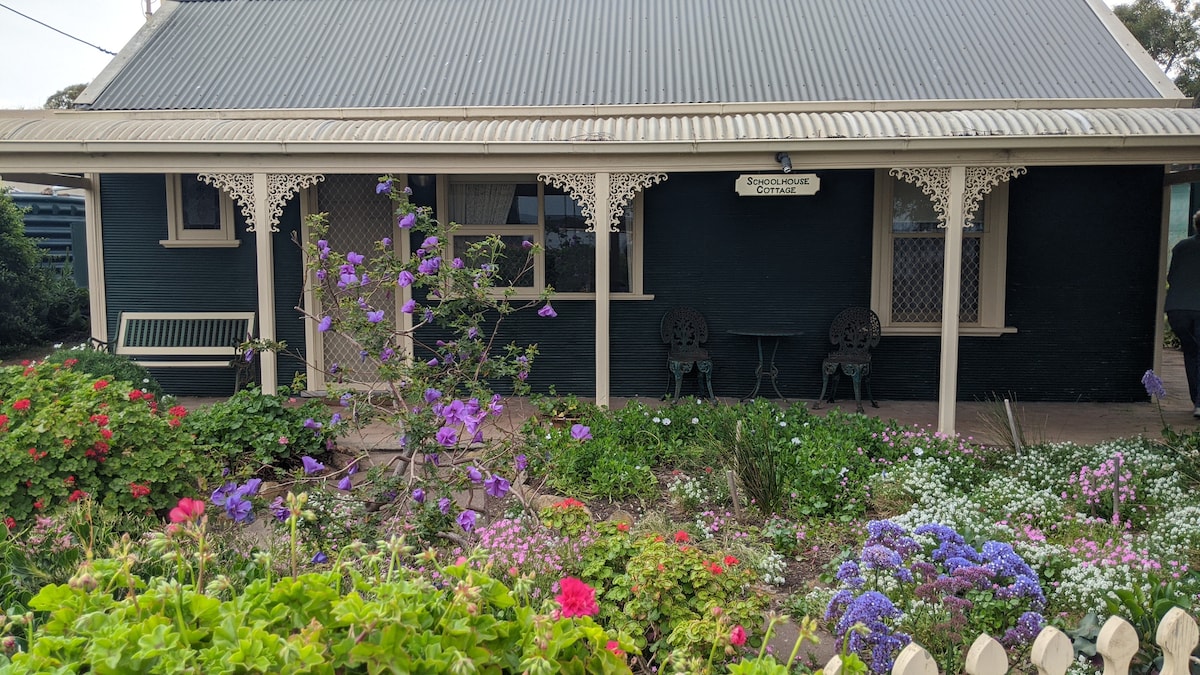 A well-preserved cottage features a charming front garden filled with vibrant flowers. Intricate timber details adorn the porch, which includes a bench and two seated areas. The front of the cottage is framed by green foliage, enhancing the inviting appearance of the home.