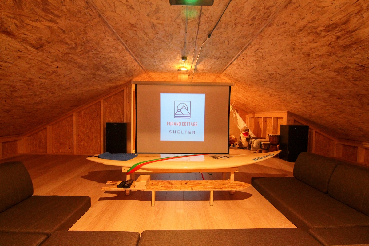 The attic space features a large screen for presentations, with a surfboard resting against a low table. Two spacious sofas are arranged for comfortable seating, providing a cozy area for gatherings or entertainment.