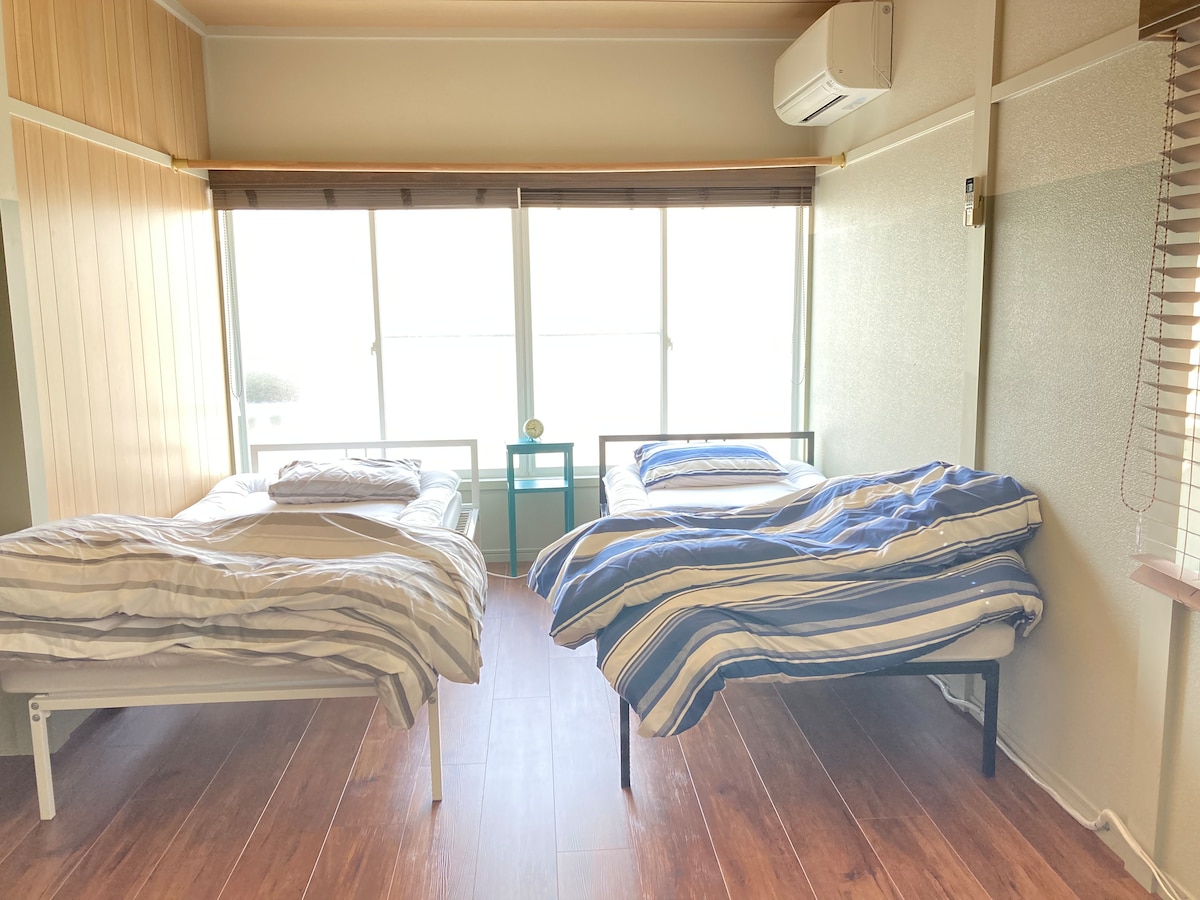 A bright sleeping area features two metal frame beds with simple bedding, complemented by natural light streaming through large windows. The room includes a green nightstand positioned between the beds, enhancing the airy and open atmosphere.