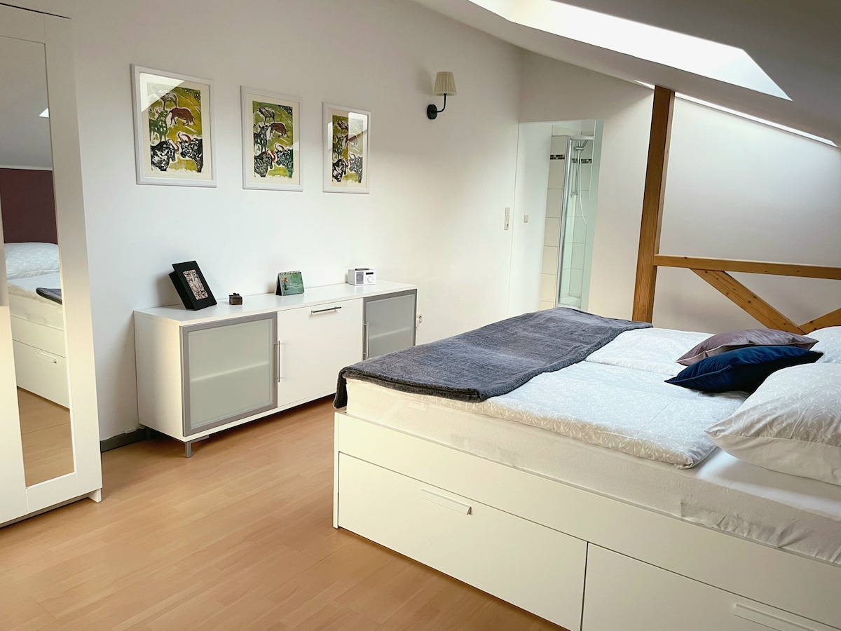 A bright sleeping area features a spacious bed dressed in crisp white linens and accented with dark pillows. Light wood flooring complements a modern dresser and console, while framed artwork adds a touch of color to the room. A glass door leads to an adjoining bathroom.