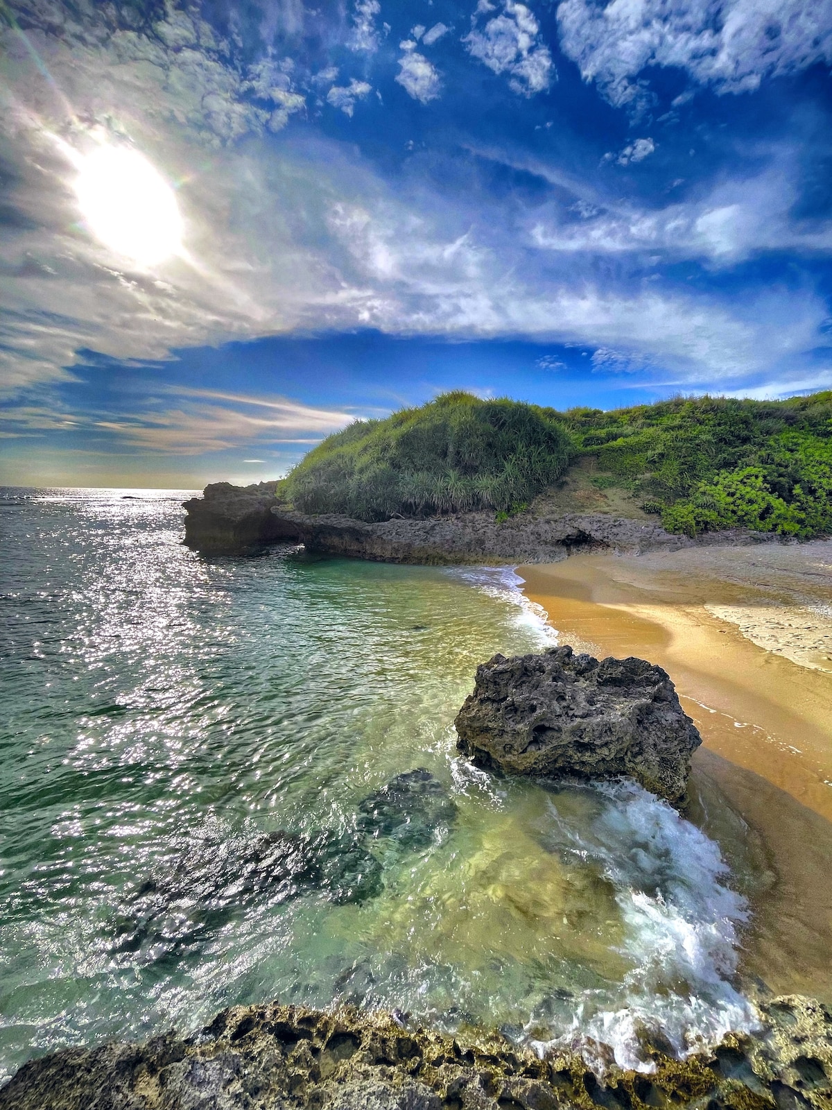 A serene beach scene displays gentle waves lapping at the shoreline, with a rocky formation partially submerged in the foreground. Lush greenery adorns the cliffs in the background, while a vibrant sky with scattered clouds is illuminated by the sun.