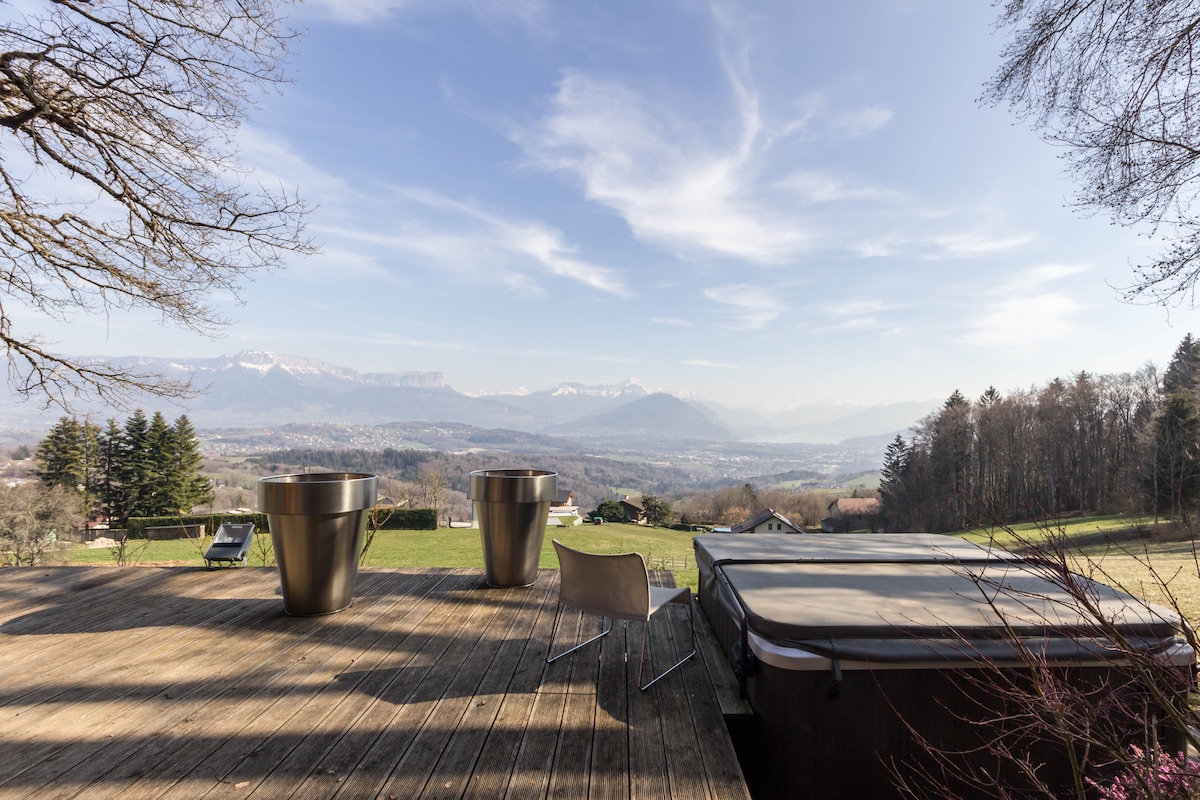A spacious wooden terrace is shown, featuring two modern metal planters and a comfortable chair overlooking a valley. The jacuzzi is positioned nearby, with distant mountain ranges visible under a clear sky, creating a serene outdoor space surrounded by nature.