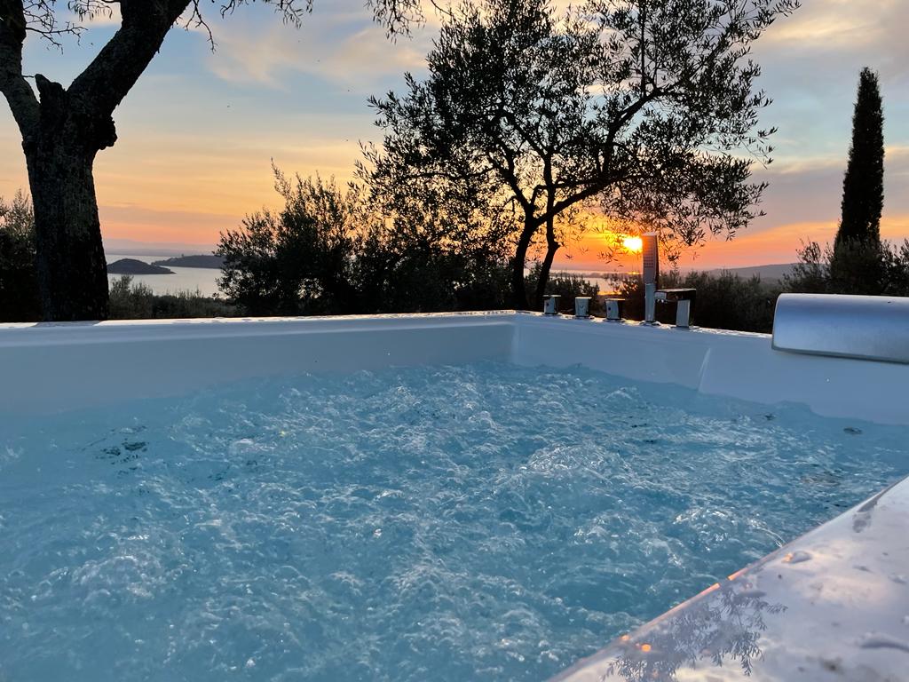 An outdoor hot tub is filled with bubbling water, framed by olive trees. A vibrant sunset casts warm colors across the sky, while the distant islands of Lake Trasimeno are visible in the background.