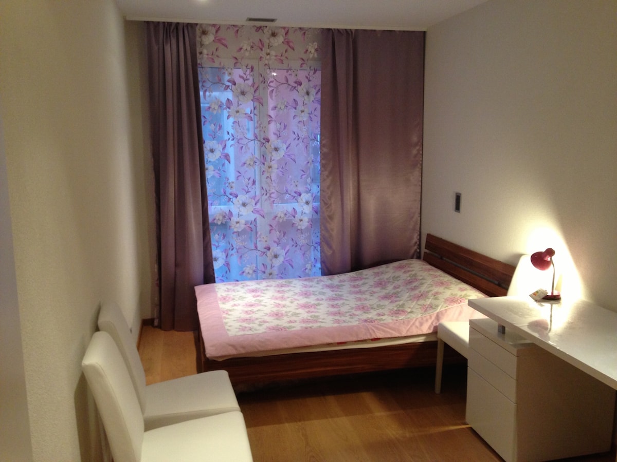 A relaxing bedroom features soft, muted tones, with a spacious bed adorned with a floral-patterned blanket. Two white chairs are positioned near a marble desk, while light filters through sheer floral curtains, creating a calm ambiance.