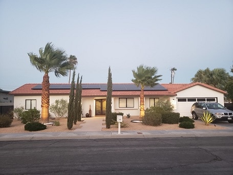 A modern single-story home features a welcoming entry with a landscaped front yard adorned with palm trees and cacti. Solar panels line the roof, and a driveway accommodates a parked vehicle. The exterior is illuminated softly, showcasing the building's clean lines and inviting appearance.